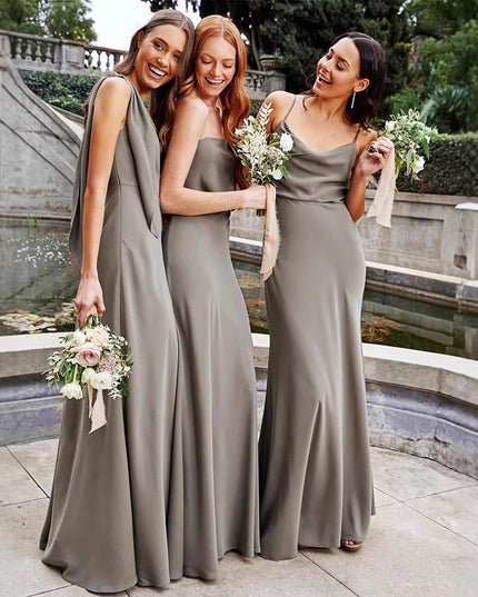 Three women in long, light gray dresses standing outdoors near a stone wall and water feature.