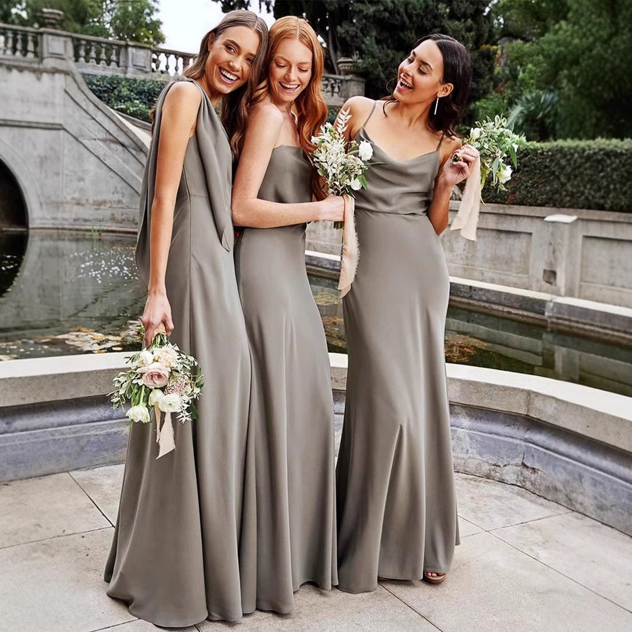 Three women in long, light gray dresses standing outdoors near a stone wall and water feature.