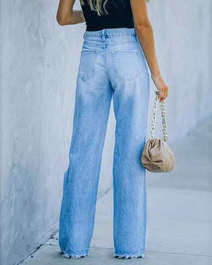 Person wearing light blue jeans and holding a beige handbag against a gray wall.