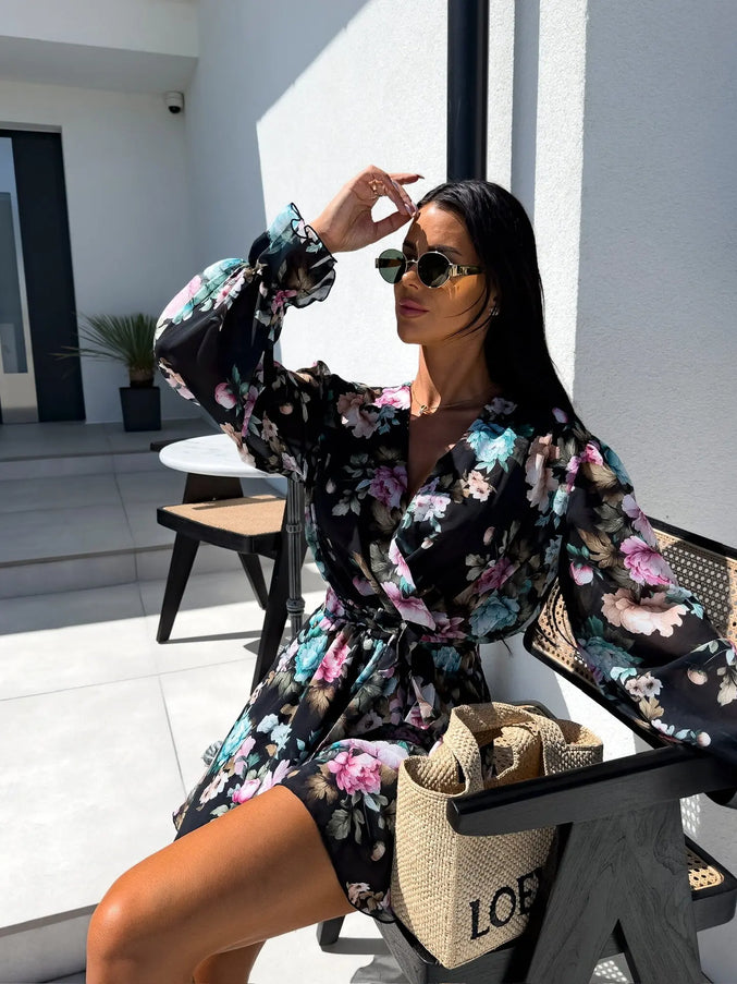Woman in a floral dress sitting outdoors with sunglasses on, holding a bag.