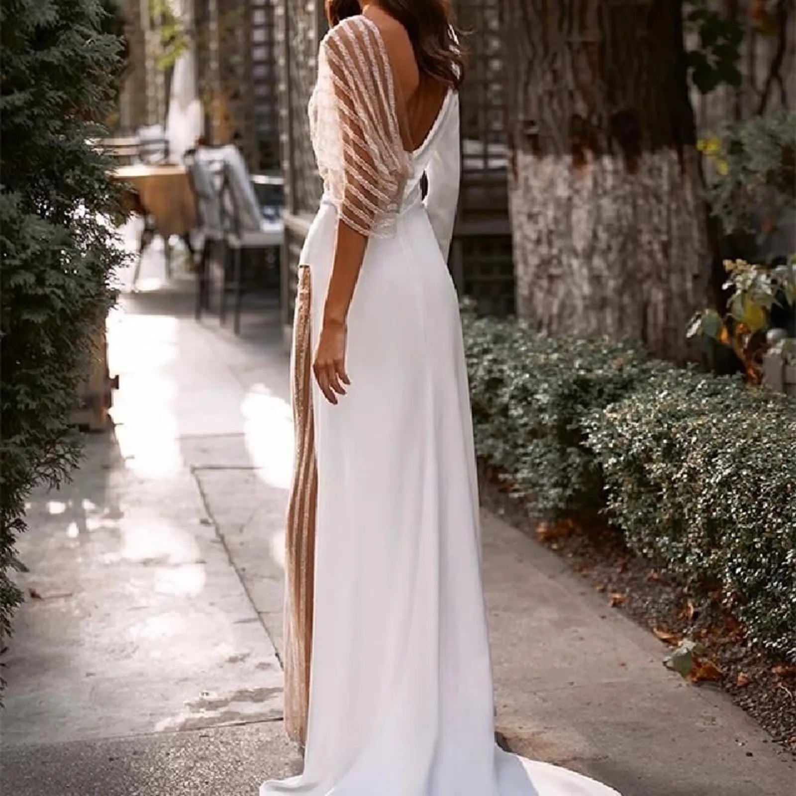 Elegant white dress with sheer lace sleeves, showcasing a deep V-back in a beautiful outdoor setting.