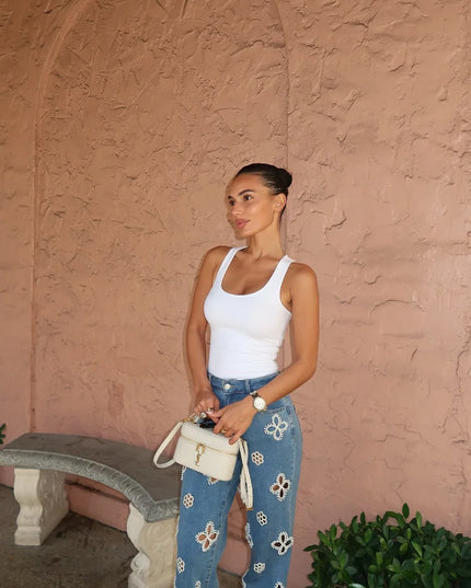 Woman in a white tank top and blue floral pants holding a white handbag against a pink wall.