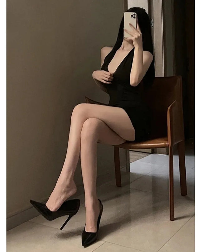 Woman wearing a black halter backless mini dress, sitting on a chair with high heels.