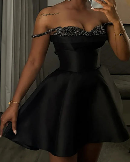 Woman wearing a black off-shoulder dress with embellishments, standing indoors.