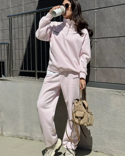 Person wearing a pink tracksuit with a beige backpack against a gray wall.