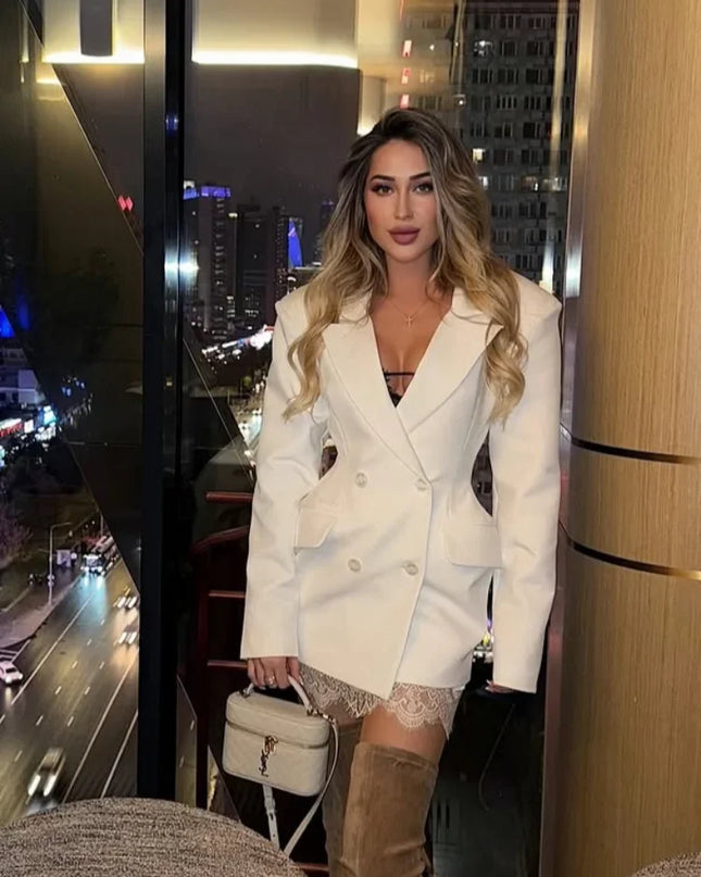 Woman in a white blazer and beige boots standing in an urban setting with city lights.