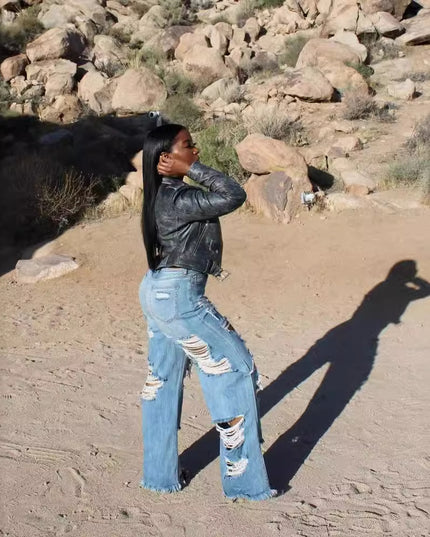 Person wearing a black leather jacket and ripped jeans standing in a desert landscape with rocks and sparse vegetation.
