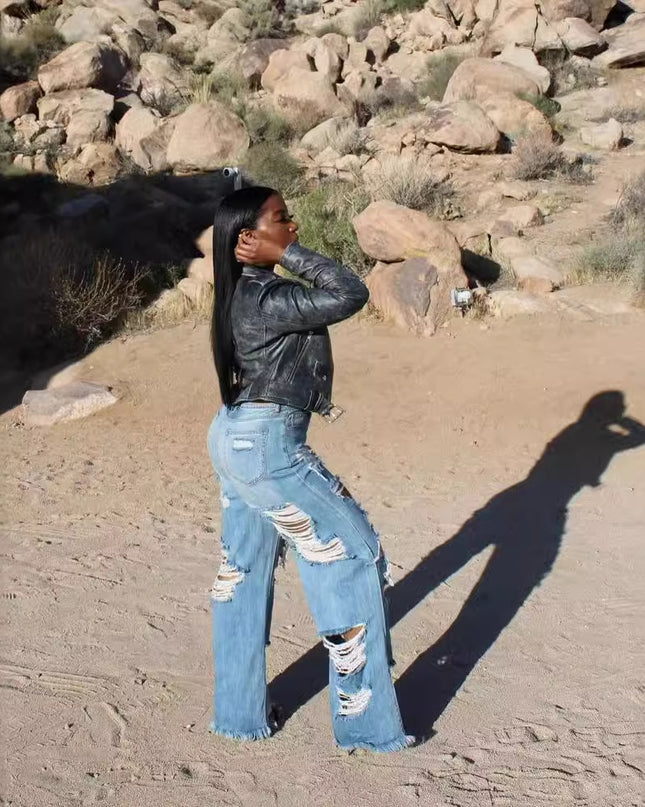 Person wearing a black leather jacket and ripped jeans standing in a desert landscape with rocks and sparse vegetation.